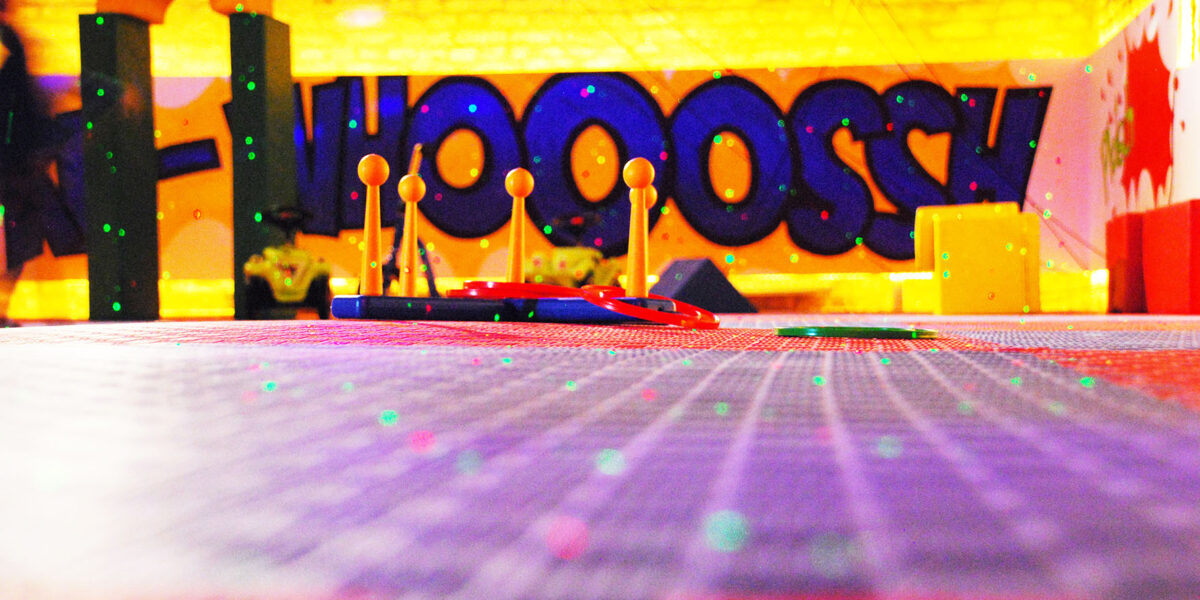 Bunter Indoor-Spielbereich mit Schwerpunkt auf Spielzeug-Hindernisparcours-Elementen, Graffiti-Stil "WHOOOSH" an der Wand und verstreuten grünen Lichtern auf dem Boden.