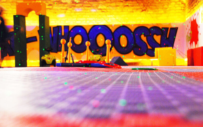 Bunter Indoor-Spielbereich mit Schwerpunkt auf Spielzeug-Hindernisparcours-Elementen, Graffiti-Stil "WHOOOSH" an der Wand und verstreuten grünen Lichtern auf dem Boden.