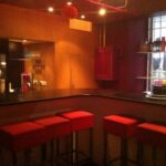 Ein schwach beleuchteter Barbereich mit einer schwarzen Arbeitsplatte, roten gepolsterten Hockern und Regalen mit Tellern und Gläsern in der Nähe eines Fensters.