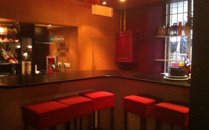 Ein schwach beleuchteter Barbereich mit einer schwarzen Arbeitsplatte, roten gepolsterten Hockern und Regalen mit Tellern und Gläsern in der Nähe eines Fensters.