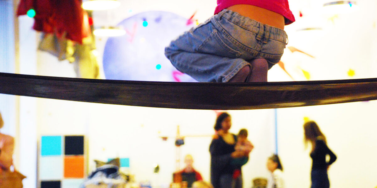 Ein Kind in Jeans und rosafarbenem Hemd balanciert auf einer Slackline in einer Halle, während im Hintergrund mehrere Erwachsene und Kinder zu sehen sind.