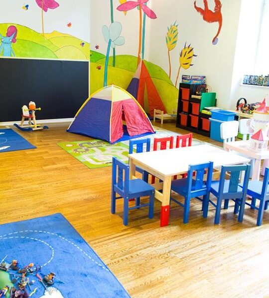 Buntes Kinderspielzimmer mit kleinen Tischen und Stühlen, Spielzeug, einem Spielzelt und farbenfrohen Wandmalereien mit Tieren und Pflanzen.