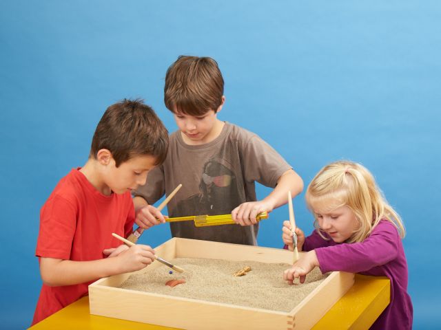 Vor einem schlichten blauen Hintergrund graben drei Kinder mit Werkzeugen in einer mit Sand gefüllten Tischkiste und suchen nach Gegenständen.