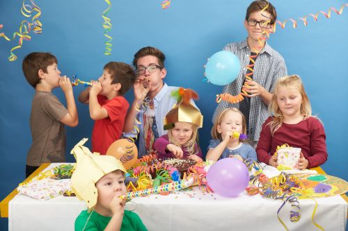 Sieben Kinder sitzen und stehen bei einer Geburtstagsparty um einen dekorierten Tisch, tragen Partyhüte, blasen Krachmacher und halten Luftballons in der Hand.