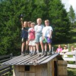 Fünf Kinder stehen auf dem Dach eines kleinen hölzernen Spielhauses, während andere Kinder und ein Erwachsener in der Nähe in einem grasbewachsenen, bewaldeten Außenbereich stehen.