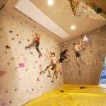 Fünf Personen klettern in einer Boulderwand mit bunten Griffen. Der Boden ist mit einer gelben Sicherheitsmatte bedeckt, im Hintergrund sind große Fenster zu sehen.