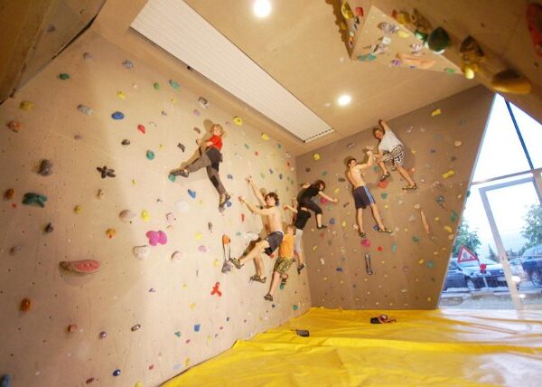 Fünf Personen klettern in einer Boulderwand mit bunten Griffen. Der Boden ist mit einer gelben Sicherheitsmatte bedeckt, im Hintergrund sind große Fenster zu sehen.