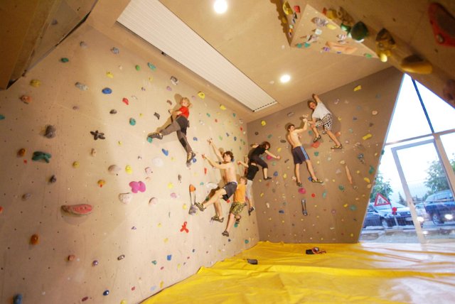 Fünf Personen klettern in einer Boulderwand mit bunten Griffen. Der Boden ist mit einer gelben Sicherheitsmatte bedeckt, im Hintergrund sind große Fenster zu sehen.