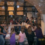 Eine Gruppe von Menschen und Kindern sitzt in einem rustikalen Raum um Tische herum und nimmt vermutlich an einem Workshop oder einer Gruppenaktivität teil. Lagerregale und gewölbte Decke sind sichtbar.