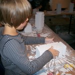 Ein Kind in einem gestreiften Hemd arbeitet mit weißen Bauklötzen an einem mit Bastelbedarf bedeckten Tisch.
