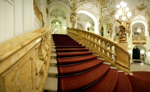 Große Treppe mit rotem Teppich, verziertem Geländer und dekorativen Goldakzenten in einem eleganten, historischen Gebäude mit hohen Decken und aufwendiger Beleuchtung.