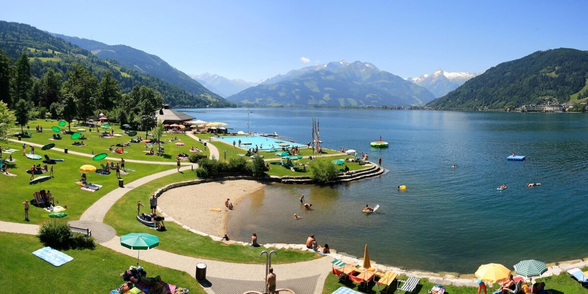 An einem sonnigen Tag entspannen sich Menschen auf einer grasbewachsenen Uferfläche mit Sonnenschirmen und schwimmen in einem großen, von Bergen umgebenen See.