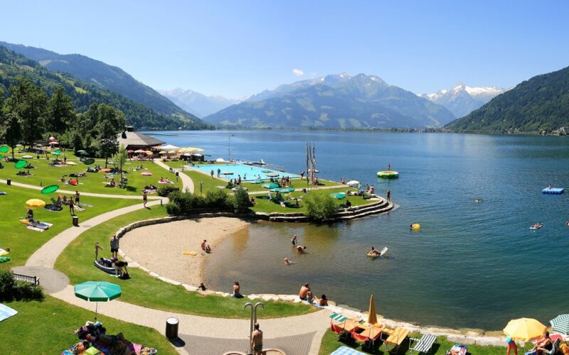 An einem sonnigen Tag entspannen sich Menschen auf einer grasbewachsenen Uferfläche mit Sonnenschirmen und schwimmen in einem großen, von Bergen umgebenen See.
