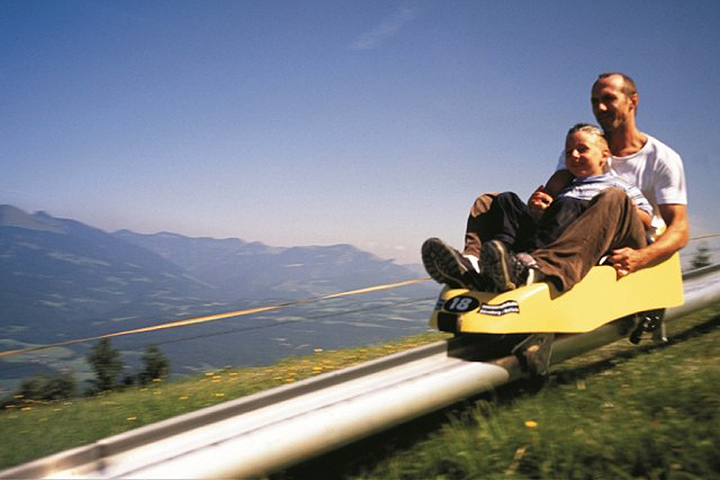 Ein Erwachsener und ein Kind fahren mit einer gelben Alpenachterbahn einen Berg hinunter, mit malerischen Hügeln und einem klaren Himmel im Hintergrund.