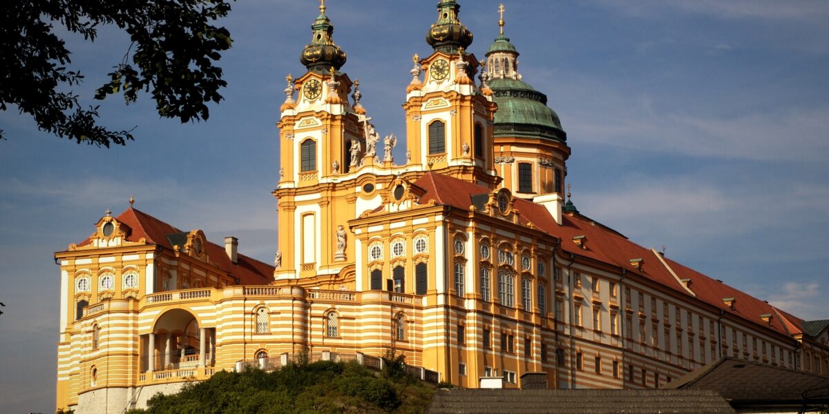 Das barocke Stift Melk in Österreich mit seinen verschnörkelten Türmen, der goldgelben Fassade und dem Kuppeldach liegt auf einem Hügel vor blauem Himmel.