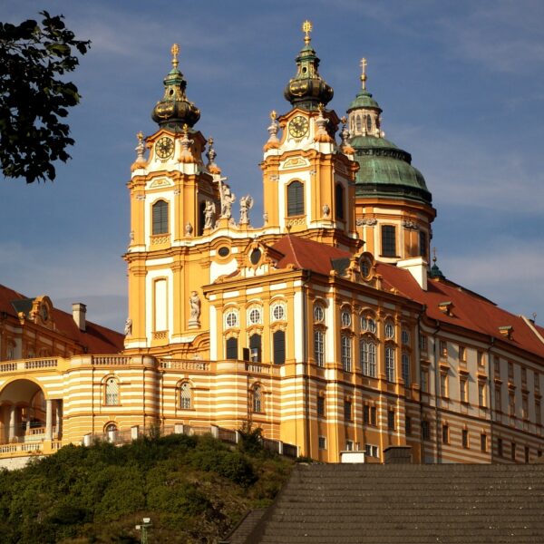 Das barocke Stift Melk in Österreich mit seinen verschnörkelten Türmen, der goldgelben Fassade und dem Kuppeldach liegt auf einem Hügel vor blauem Himmel.