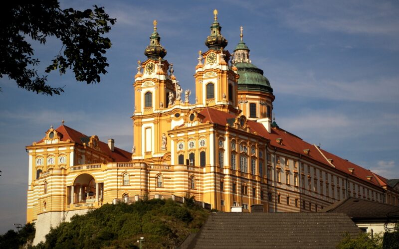 Das barocke Stift Melk in Österreich mit seinen verschnörkelten Türmen, der goldgelben Fassade und dem Kuppeldach liegt auf einem Hügel vor blauem Himmel.