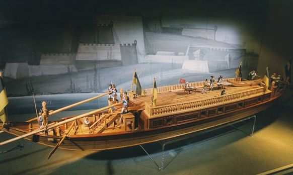 Ein detailliertes Modell eines antiken hölzernen Kriegsschiffs mit menschlichen Miniaturfiguren, Rudern und Flaggen wird in einer Museumsumgebung vor einem Wandhintergrund ausgestellt.