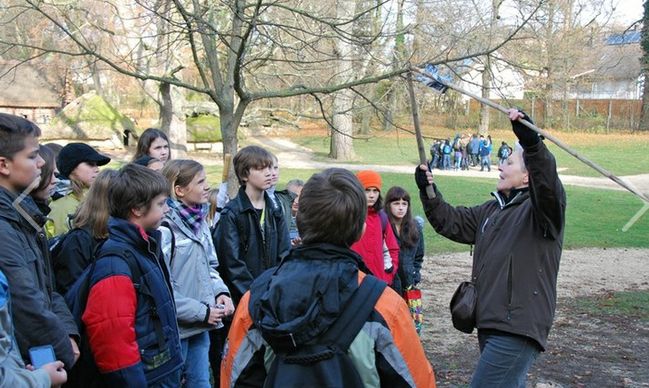 Eine Gruppe Kinder steht draußen in einem Park und hört einem Erwachsenen zu, der einen langen Stock hält und beim Sprechen gestikuliert.