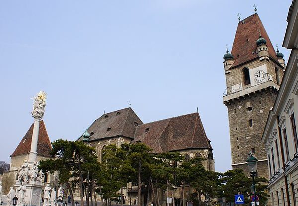 Historische Steingebäude mit einem hohen Uhrenturm, einem kunstvollen Denkmal und Bäumen davor, unter einem klaren Himmel.