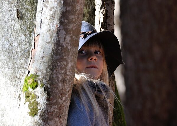 Ein junges Mädchen mit breitkrempigem Hut steht zwischen Baumstämmen, teilweise im Schatten, und blickt leicht zur Seite.