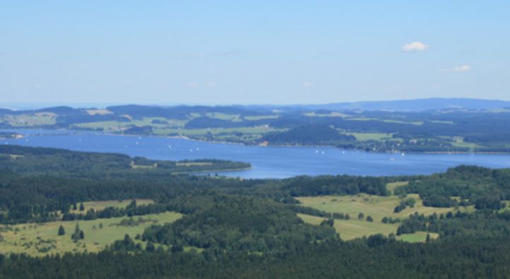 Ein breiter Fluss oder See mit umgebenden grünen Wäldern und Feldern unter einem klaren blauen Himmel, von oben betrachtet.