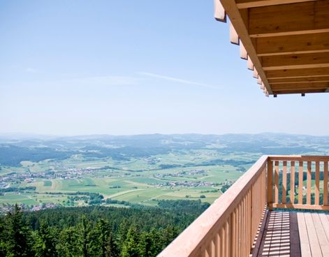 Von einem Holzbalkon mit Geländer und Dachüberstand blickt man auf ein weites, grünes Tal mit verstreuten Gebäuden, Bäumen und fernen Hügeln unter einem klaren blauen Himmel.