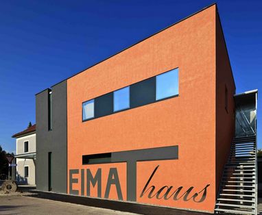 Modernes zweistöckiges Gebäude mit orange-dunkelgrauer Fassade, großen Fenstern, Außentreppe und dem Schriftzug „EIMAT haus“ an der Vorderwand.