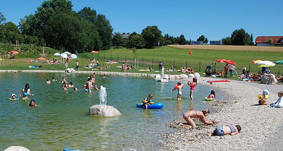 Menschen schwimmen und spielen in einem natürlichen Freibad mit Kiesufer. Andere entspannen an einem sonnigen Tag auf der Wiese mit Sonnenschirmen.