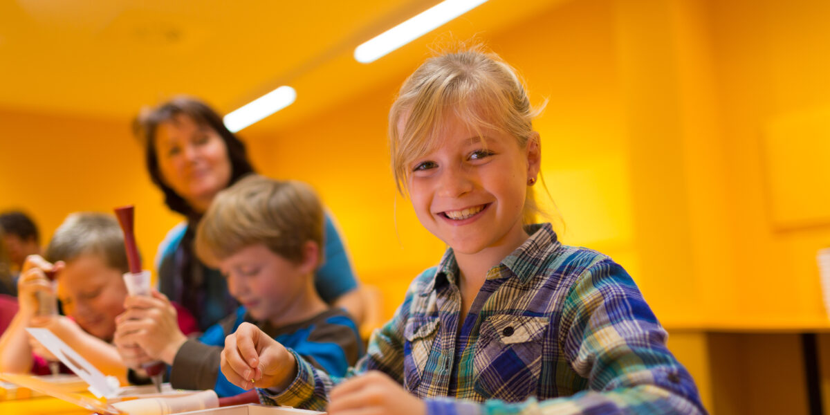 Ein lächelndes Mädchen arbeitet an einem Tisch mit anderen Kindern an einem Bastelprojekt, während ein Erwachsener im Hintergrund in einem hell erleuchteten gelben Raum steht.