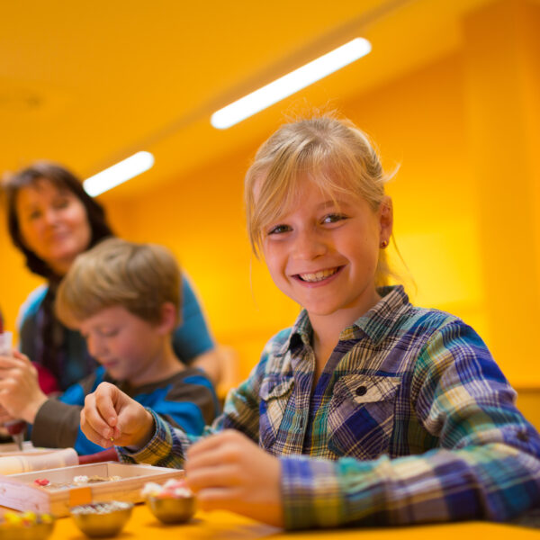 Ein lächelndes Mädchen arbeitet an einem Tisch mit anderen Kindern an einem Bastelprojekt, während ein Erwachsener im Hintergrund in einem hell erleuchteten gelben Raum steht.