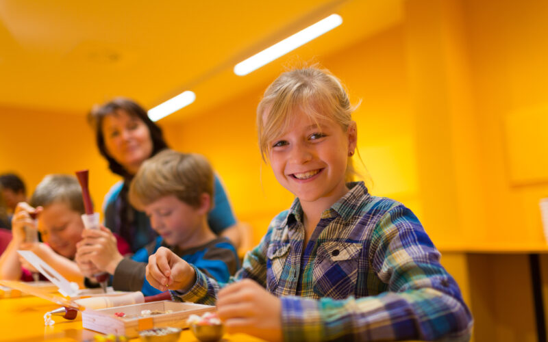 Ein lächelndes Mädchen arbeitet an einem Tisch mit anderen Kindern an einem Bastelprojekt, während ein Erwachsener im Hintergrund in einem hell erleuchteten gelben Raum steht.