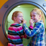 Zwei junge Mädchen stehen vor einem runden Fenster in einer gelben Wand, lächeln und essen Snacks.