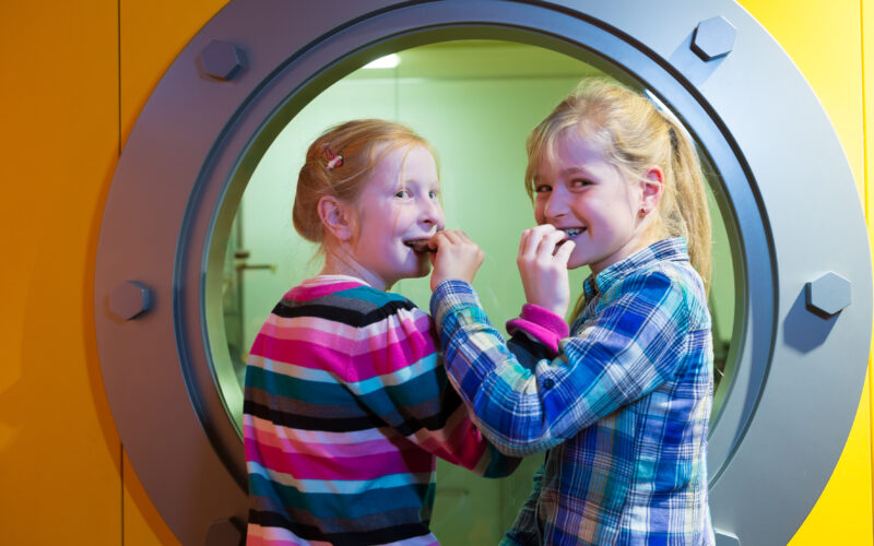 Zwei junge Mädchen stehen vor einem runden Fenster in einer gelben Wand, lächeln und essen Snacks.