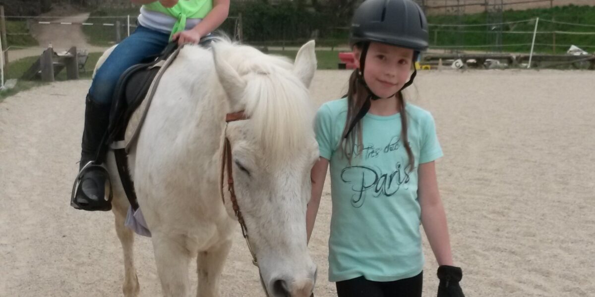 Zwei junge Mädchen mit Helmen, von denen die eine ein weißes Pferd reitet und die andere es an den Zügeln führt, gehen gemeinsam auf einem Reitplatz im Freien.
