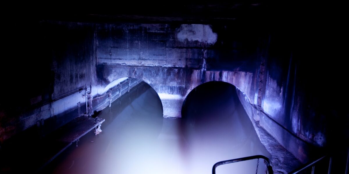 Zwei große gewölbte Tunnel, die teilweise mit Wasser überflutet sind und von einer hellen Lichtquelle in einem ansonsten dunklen unterirdischen Raum beleuchtet werden.