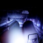 Zwei große gewölbte Tunnel, die teilweise mit Wasser überflutet sind und von einer hellen Lichtquelle in einem ansonsten dunklen unterirdischen Raum beleuchtet werden.