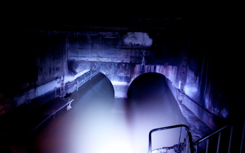 Zwei große gewölbte Tunnel, die teilweise mit Wasser überflutet sind und von einer hellen Lichtquelle in einem ansonsten dunklen unterirdischen Raum beleuchtet werden.