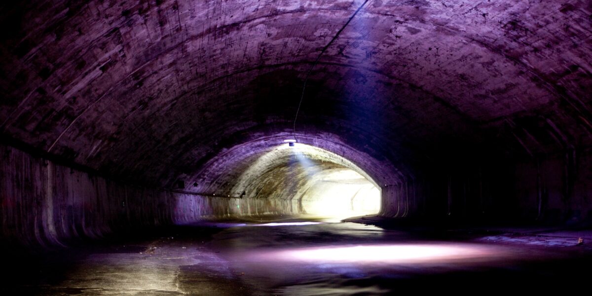 Ein Betontunnel mit gewölbter Decke wird schwach durch natürliches Licht beleuchtet, das durch eine kreisförmige Öffnung darüber und den Tunnelausgang im Hintergrund einfällt.
