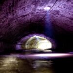 Ein Betontunnel mit gewölbter Decke wird schwach durch natürliches Licht beleuchtet, das durch eine kreisförmige Öffnung darüber und den Tunnelausgang im Hintergrund einfällt.