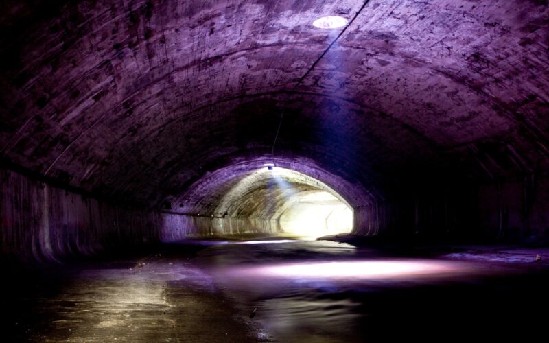 Ein Betontunnel mit gewölbter Decke wird schwach durch natürliches Licht beleuchtet, das durch eine kreisförmige Öffnung darüber und den Tunnelausgang im Hintergrund einfällt.