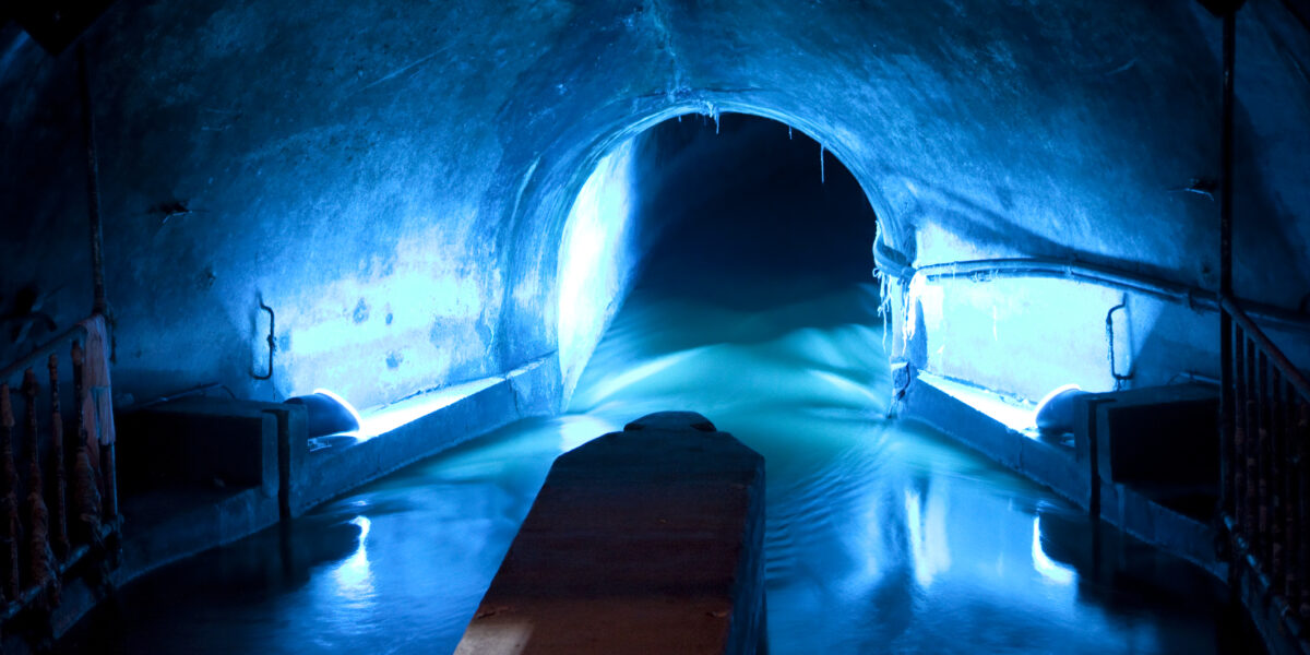 Schwach beleuchteter unterirdischer Tunnel mit blauer Beleuchtung, durch die Mitte fließendem Wasser und einer gewölbten Decke.