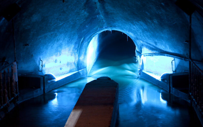 Schwach beleuchteter unterirdischer Tunnel mit blauer Beleuchtung, durch die Mitte fließendem Wasser und einer gewölbten Decke.