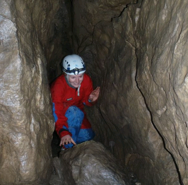 Eine Person mit Helm und roter Jacke hockt in einem engen, von Felswänden umgebenen Höhlengang.