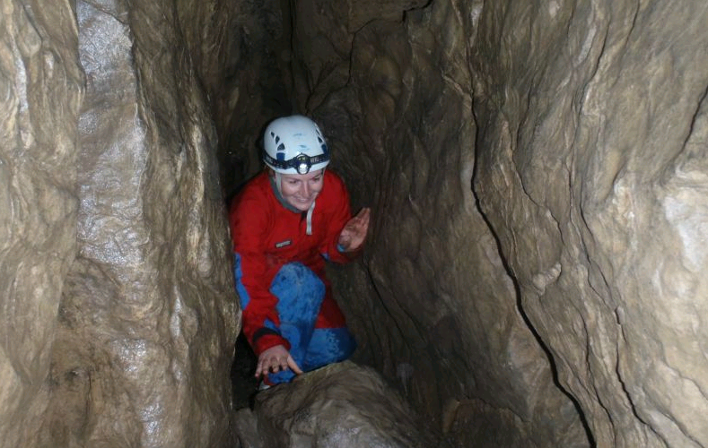 Eine Person mit Helm und roter Jacke hockt in einem engen, von Felswänden umgebenen Höhlengang.