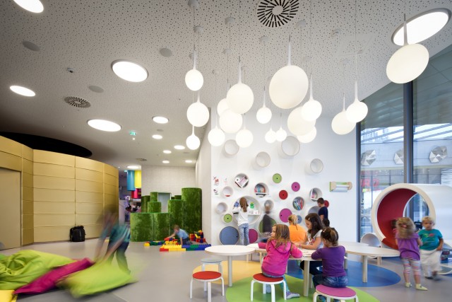 Kinder spielen und interagieren in einem hell erleuchteten, modernen Indoor-Spielzimmer mit farbenfrohen Möbeln, runden Leuchten und verspielten Wanddekorationen.