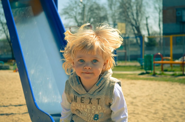 Ein kleines Kind mit blonden Haaren steht an einem sonnigen Tag neben einer blauen Rutsche auf einem Spielplatz, im Hintergrund sind Sand und Spielgeräte zu sehen.
