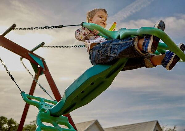 Ein kleines Kind in Jeans und Turnschuhen schaukelt draußen unter einem teilweise bewölkten Himmel auf einer grünen Plastikschaukel.