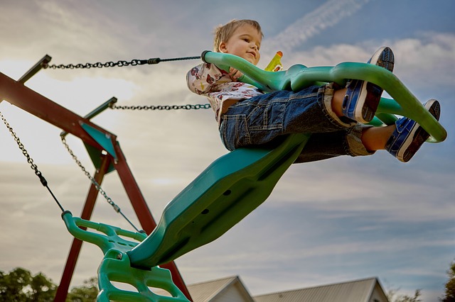 Ein kleines Kind in Jeans und Turnschuhen schaukelt draußen unter einem teilweise bewölkten Himmel auf einer grünen Plastikschaukel.