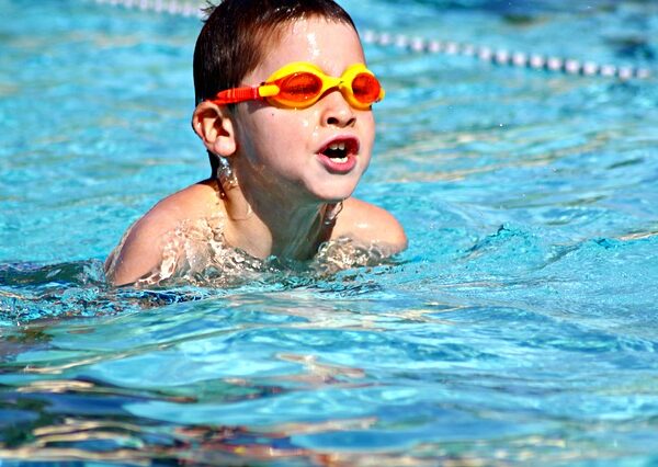 Ein kleiner Junge mit einer orangefarbenen Schwimmbrille schwimmt in einem Pool. Um ihn herum spritzt Wasser, im Hintergrund ist eine Bahnbegrenzung zu sehen.
